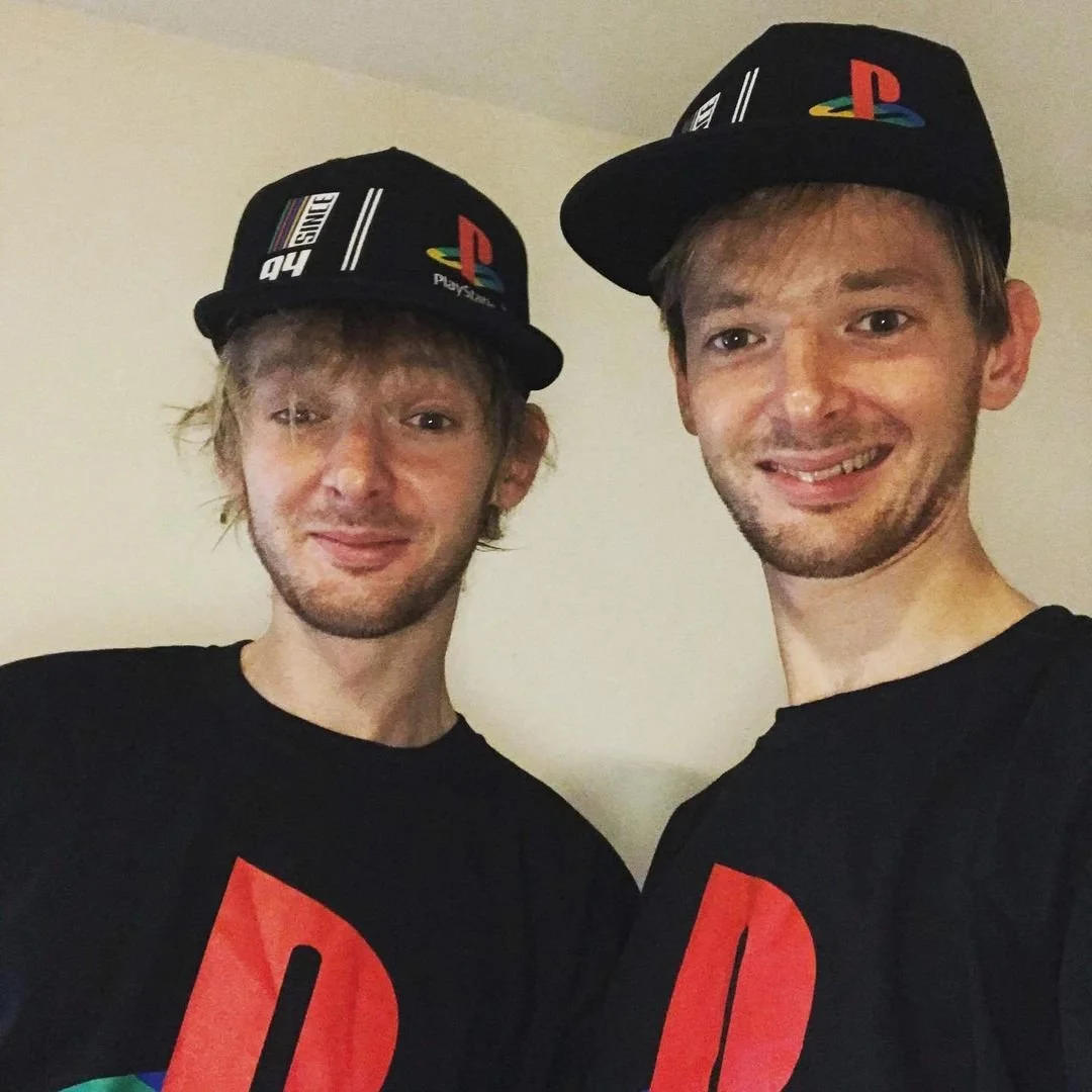 Two young men wearing black PlayStation-themed hats and matching black T-shirts with large red and blue PlayStation logos, standing together against a plain wall.