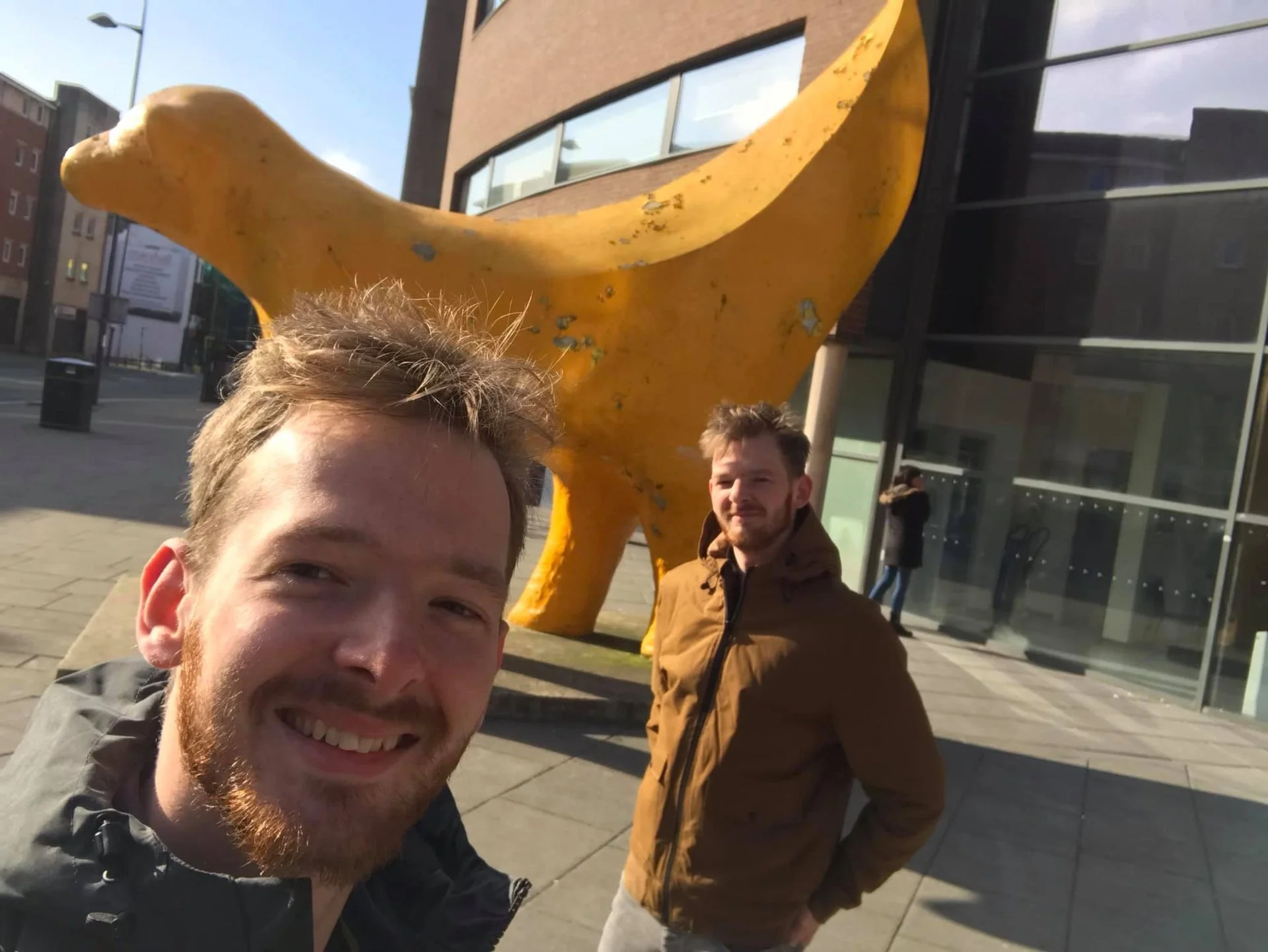 Two men taking a selfie in front of a large yellow duck sculpture on a city sidewalk.