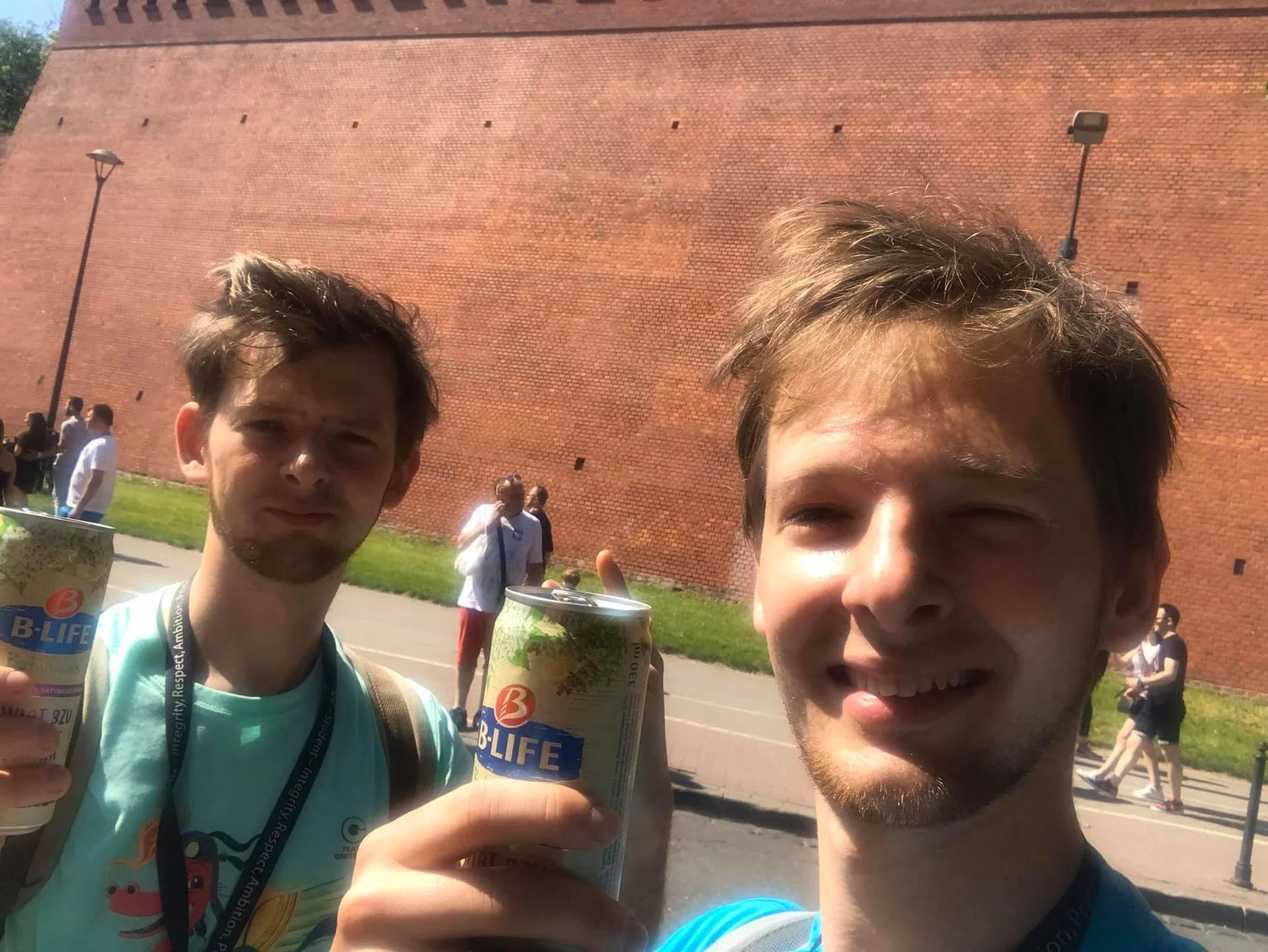 Two young men taking a selfie outdoors during daytime, holding cans of B-LIFE beverage, with a large brick wall and other people in the background.
