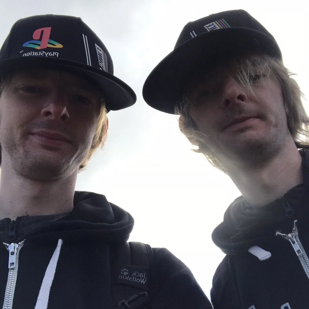 Two young men taking a selfie outdoors, wearing black caps with PlayStation and other logos, and black hoodies. The sky is cloudy in the background.