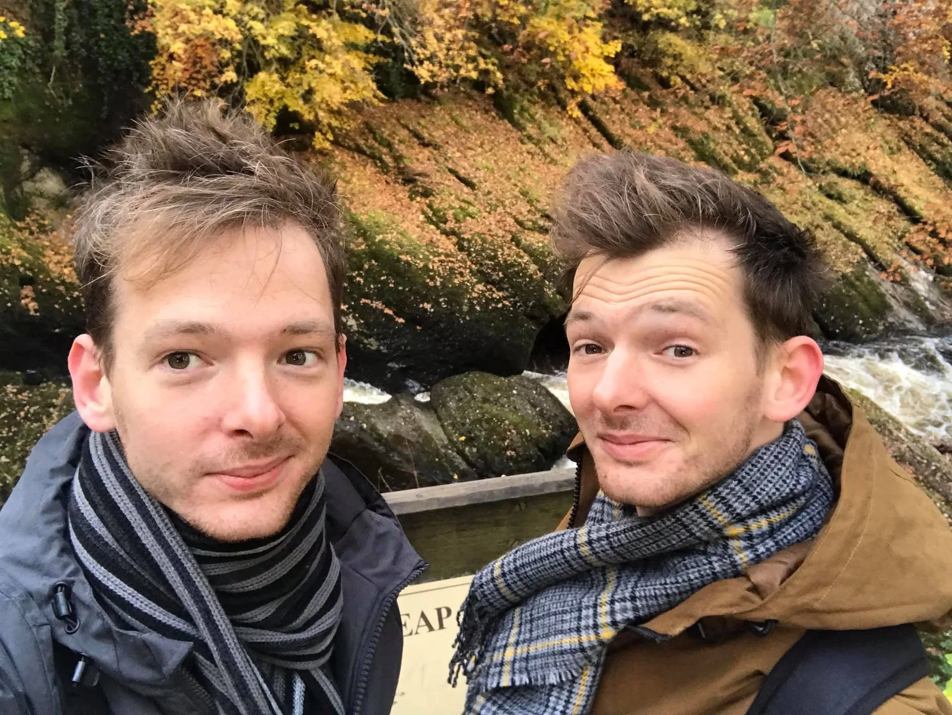 Two men taking a selfie outdoors near a river with autumn trees in the background.
