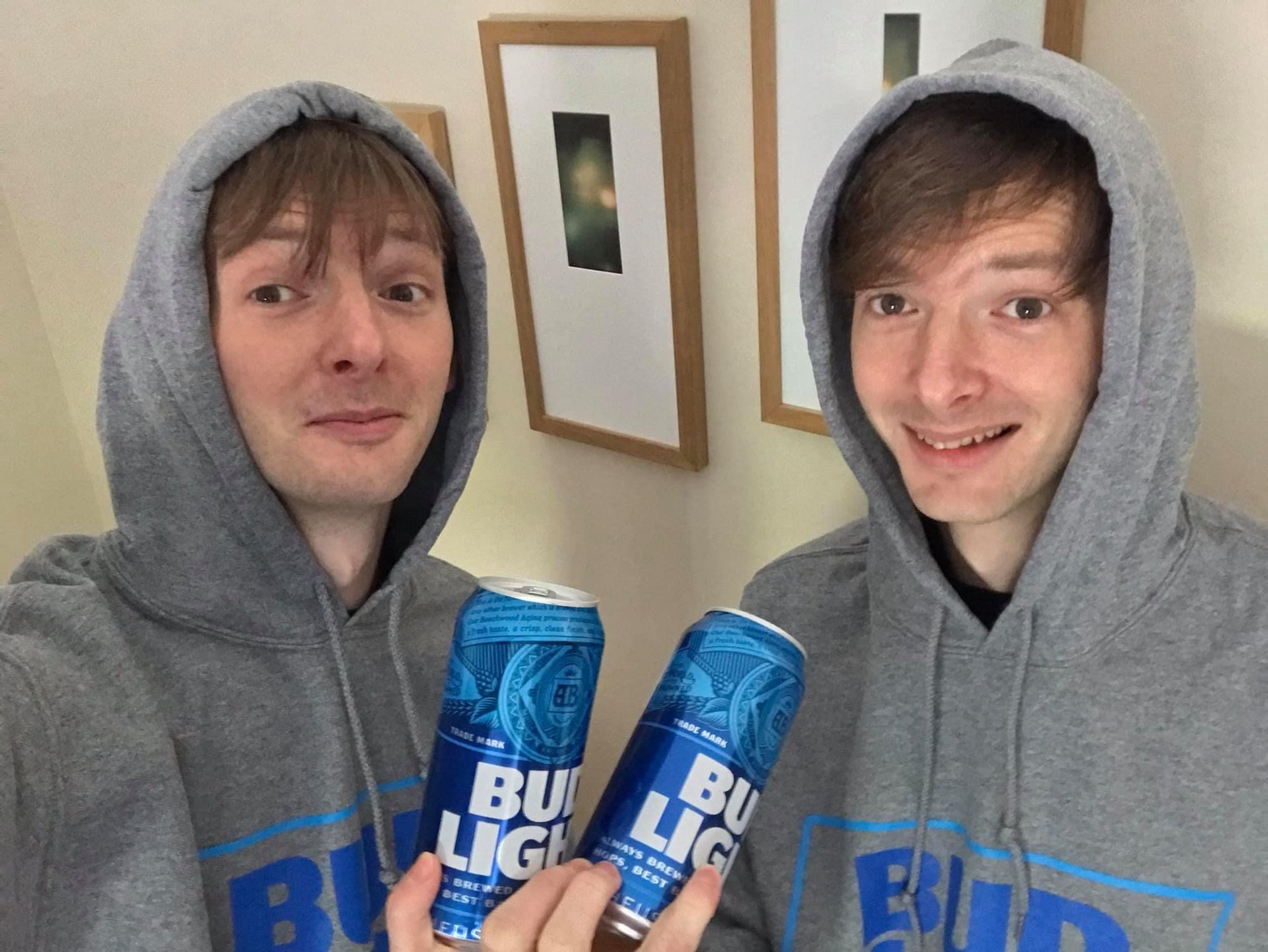 Two young men wearing gray hoodies with blue print, smiling and holding cans of Bud Light beer, taking a selfie indoors in front of a wall with framed pictures.