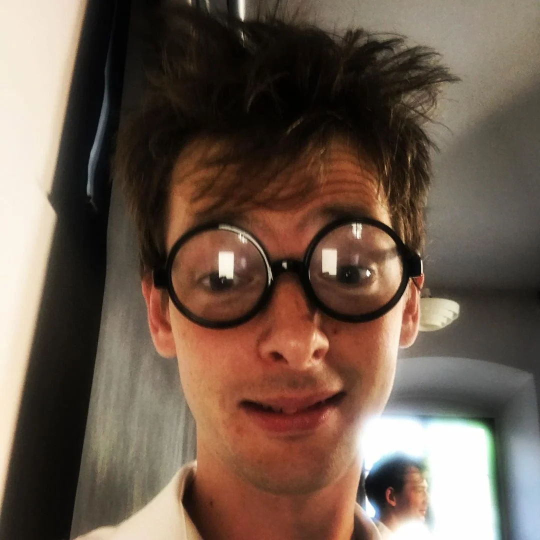 A young man with messy brown hair wearing round black glasses, smiling in an indoor setting with other people in the background.