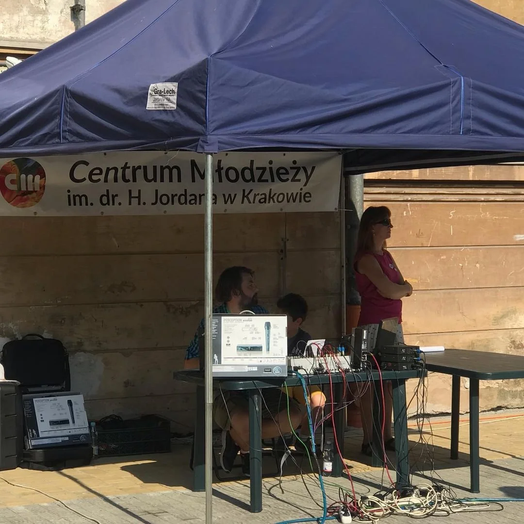 A booth with a blue canopy and a sign that reads 'Centrum Młodzieży im. dr. H. Jordana w Krakowie.' Underneath, two men are seated at a table with electronic equipment, wires, and a microphone box, with a woman standing to the side wearing sunglasses