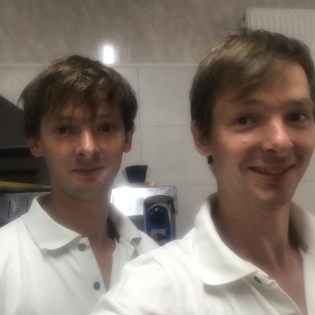 Two young men taking a selfie together indoors, both wearing white shirts, with a kitchen or bar setting in the background.