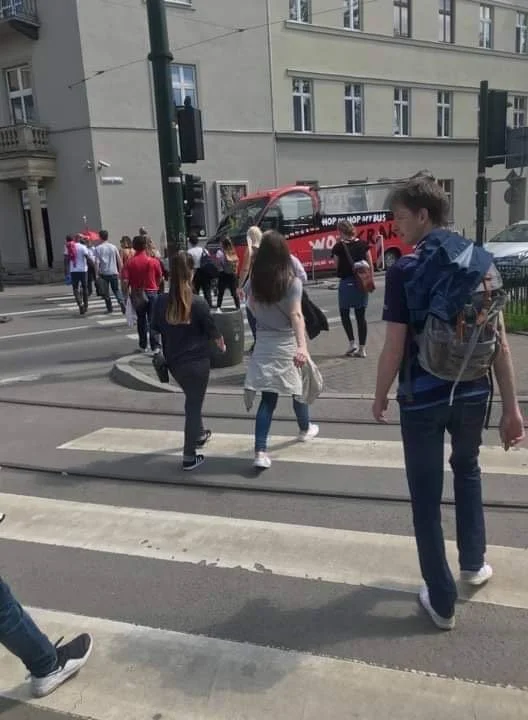 People crossing a city street at a crosswalk with a red sightseeing bus in the background.