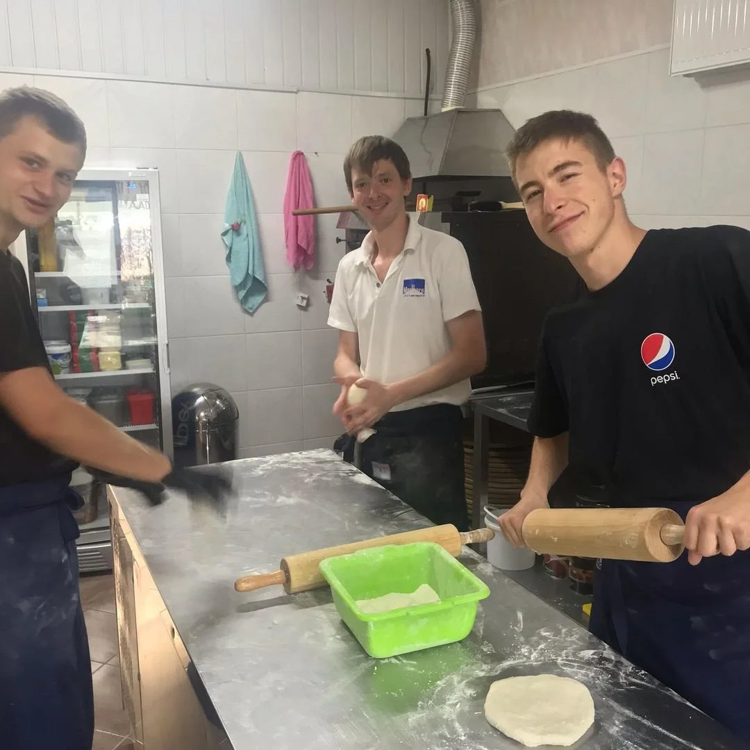 Three young men in a kitchen preparing dough, with rolling pins and flour on the table.