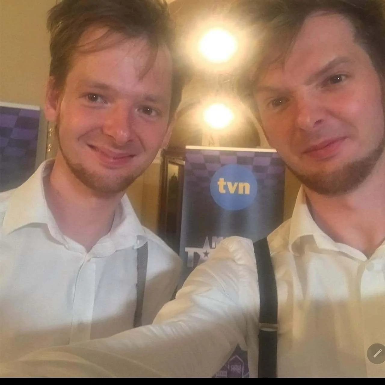 Two men in white shirts taking a selfie in front of a tvn banner at an indoor event, with warm lighting and a blurred background.