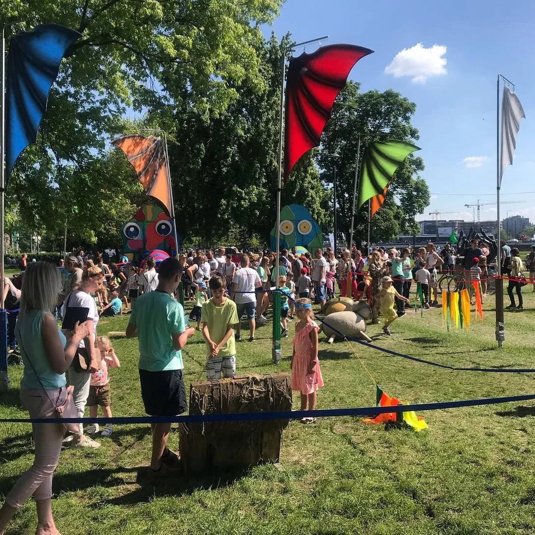 A crowd of people gathered outdoors at a festival or fair on a sunny day, with colorful flags and whimsical decorations, including large cartoonish character cutouts, in a park with green trees and a cityscape in the background.