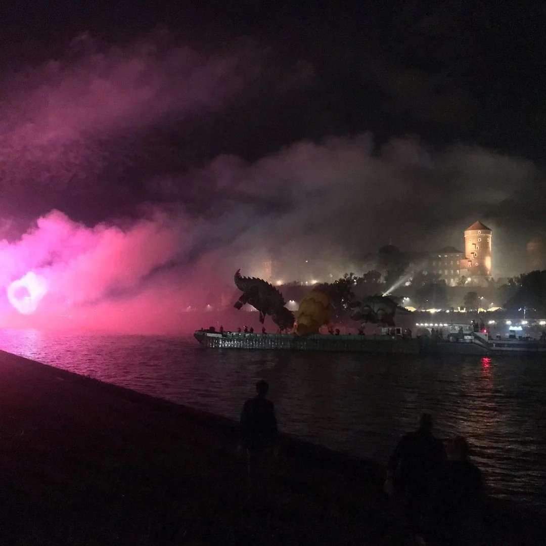Night scene with a dragon sculpture on a boat, pink and purple smoke or fog over water, and a large castle in the background illuminated with spotlights.