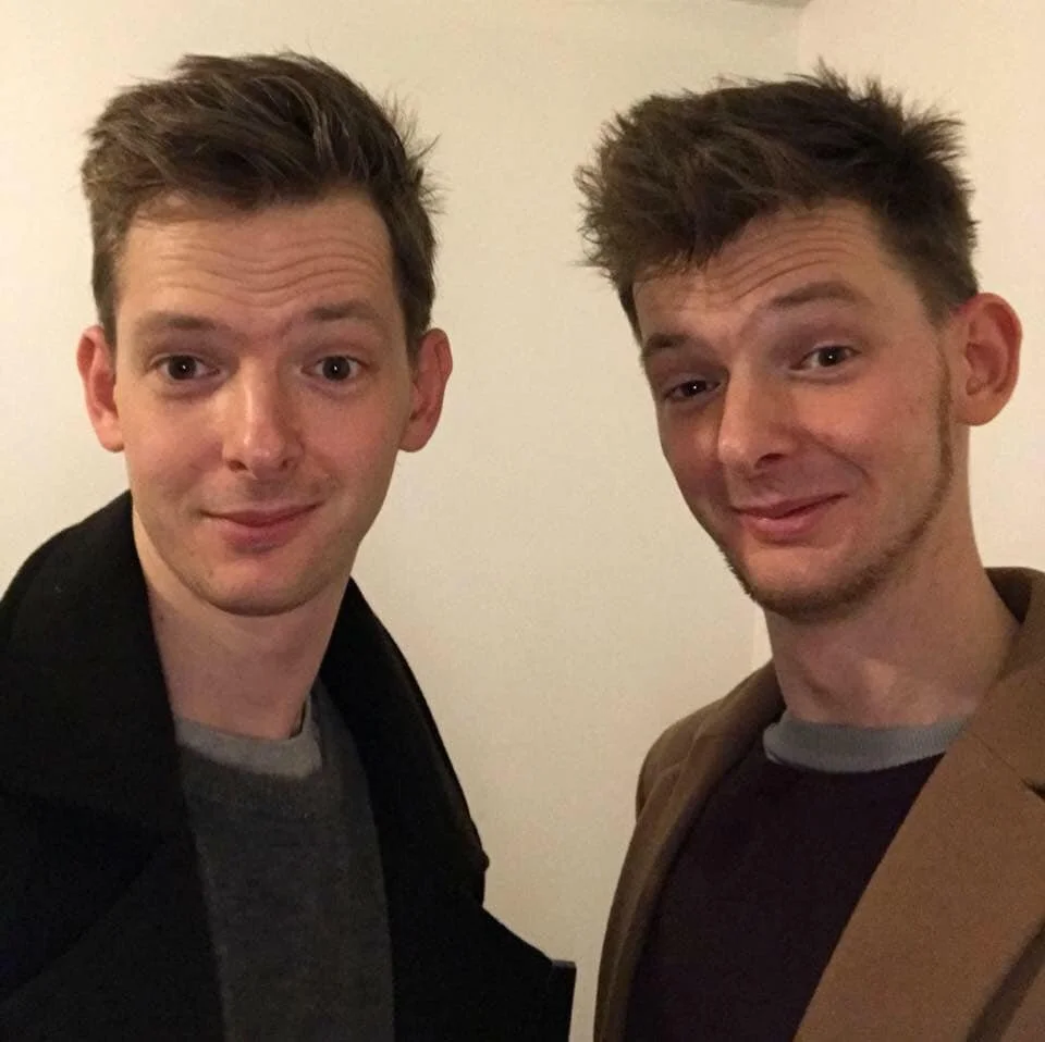 Two young men with styled hair and light skin taking a selfie together against a plain wall.