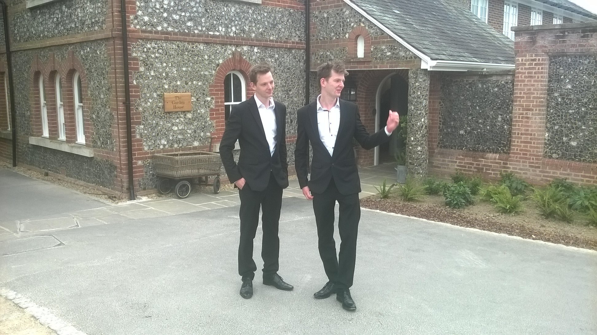 Two men in black suits and white shirts walking on a sidewalk outside a brick building with a sign that reads "The Garden House." One man is gesturing with his right hand while talking, the other is looking at him.