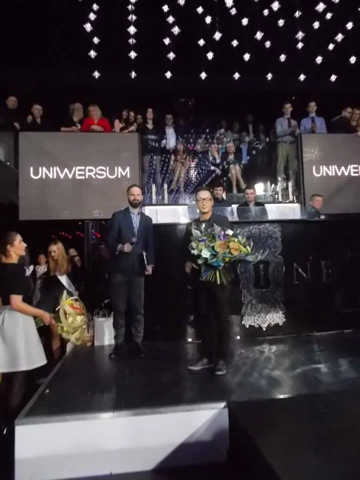 A man holding a large bouquet of flowers on stage at an event, with a man beside him holding a microphone, and a woman and children nearby. Audience members are seated above, with hanging decorative lights.