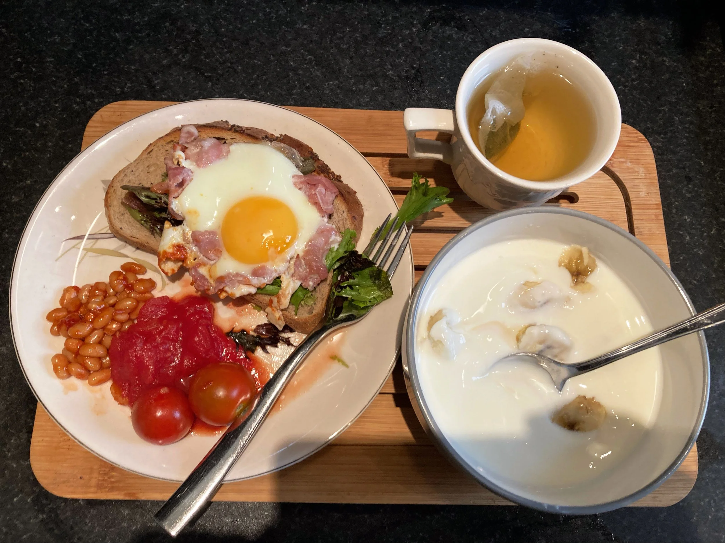 A breakfast tray with a plate of toast topped with a fried egg, bacon, and greens, alongside baked beans, cherry tomatoes, a bowl of yogurt with bananas, and a mug of tea.