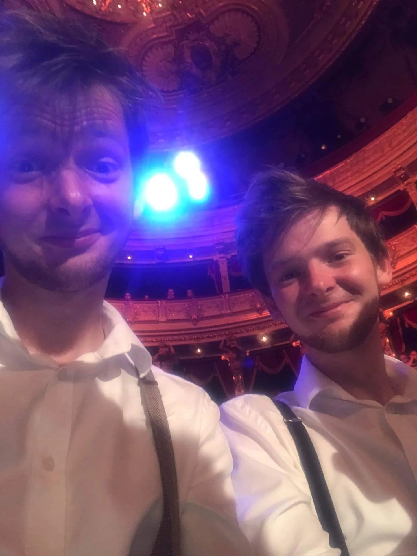 Two young men in white shirts and suspenders taking a selfie on stage inside a grand, ornately decorated theater with red and gold accents, illuminated by blue stage lights.