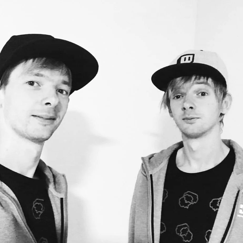 Two young men wearing baseball caps and casual hoodies, standing against a plain wall, taking a black and white selfie.