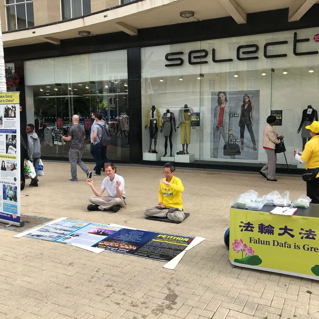 People sitting and standing outside a retail store with a protest or demonstration setup on the sidewalk. There are signs and posters on the ground and a banner with Chinese characters and the phrase 'Falun Dafa is G' on a yellow background.
