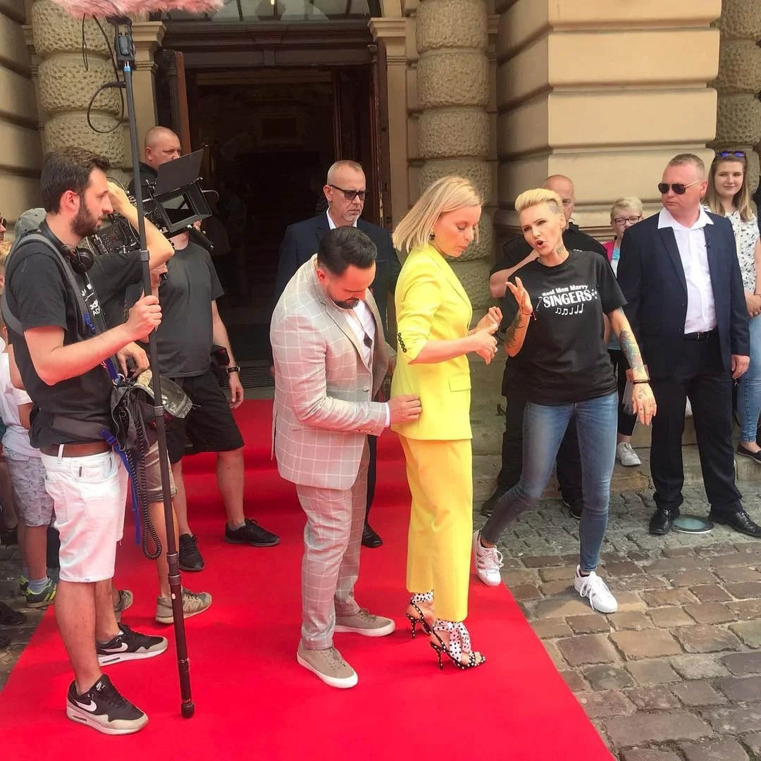 A blonde woman in a yellow suit standing on a red carpet with a group of people, some of whom are taking pictures or videos outside a historic building, during a public event or gathering.