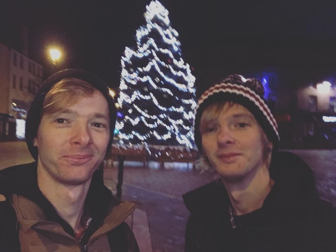 Two young men taking a selfie in front of a decorated Christmas tree at night.