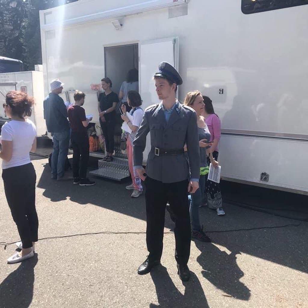A woman in a gray military-style uniform and hat standing in front of a white trailer, with people gathered around and waiting outside.