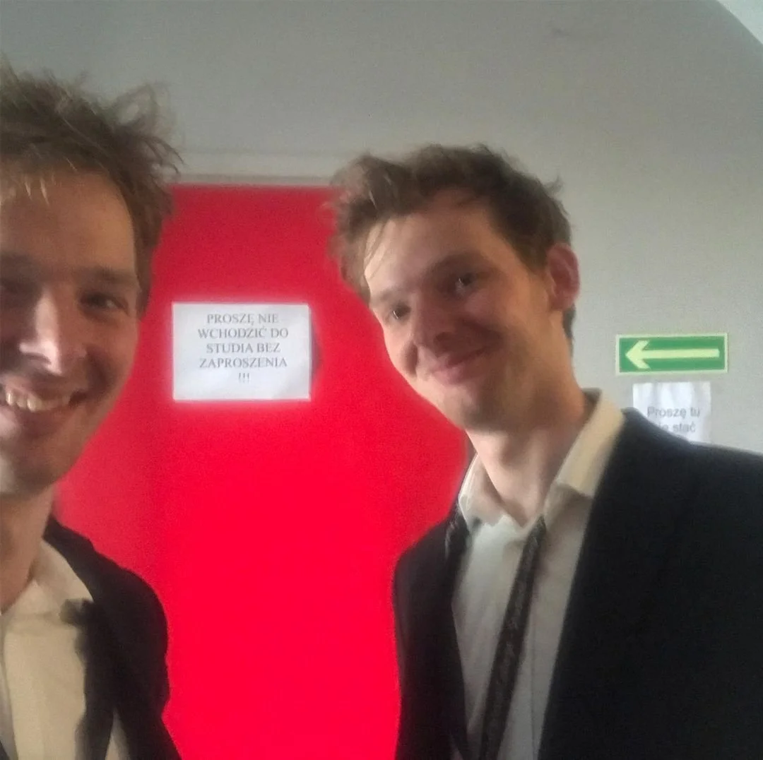 Two young men smiling and taking a selfie in front of a red door with a sign in Polish that says, 'Please do not enter the study without permission.'