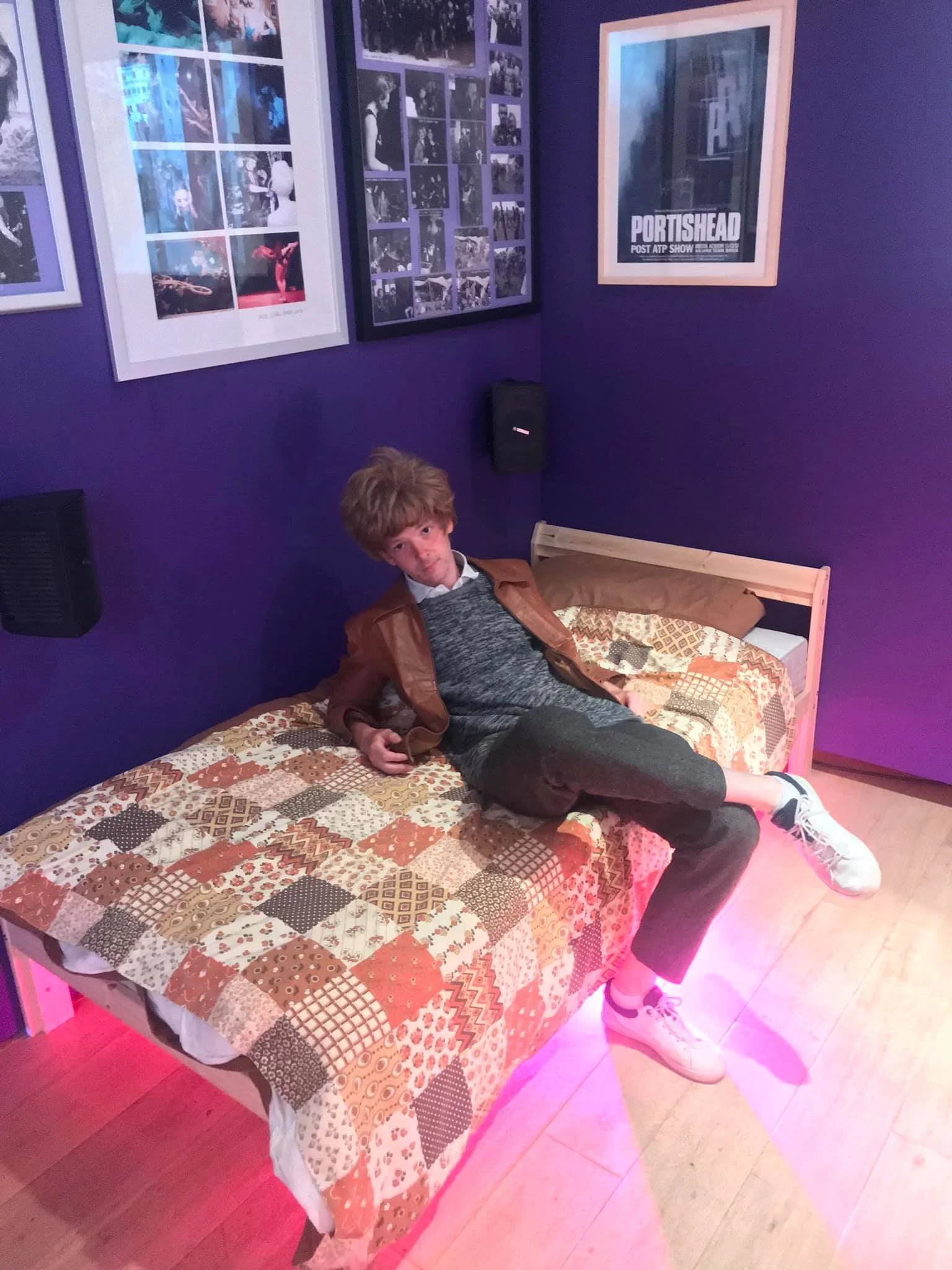 A young man with curly hair is sitting on a bed in a room with purple walls. The bed has a patchwork quilt, and there are framed posters on the wall behind him. The lighting gives a pinkish hue to the floor.