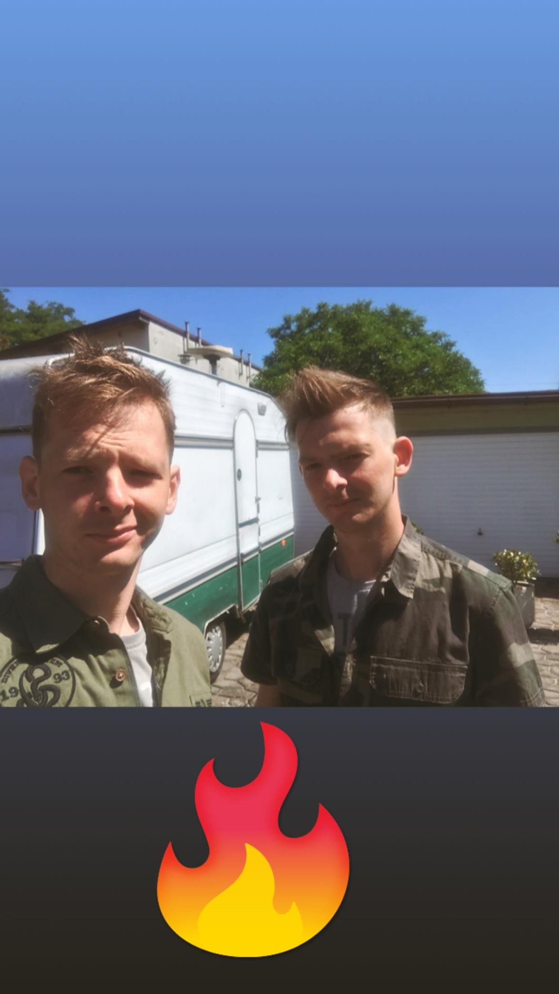 Two young men taking a selfie outdoors on a sunny day, with a trailer and a house in the background, and a fire emoji overlay on the bottom of the image.