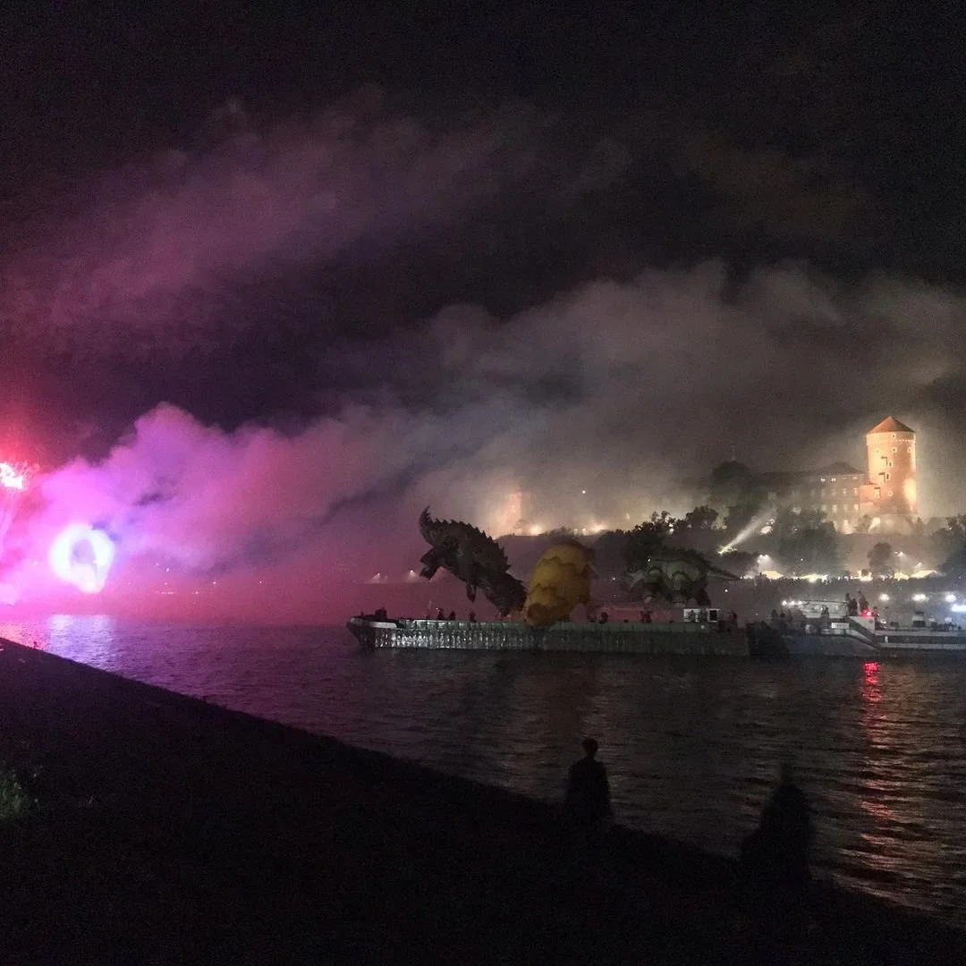 Nighttime fireworks display over water with colorful smoke, fireworks, and illuminated dragon and creature sculptures on a boat, with a castle and trees in the background.
