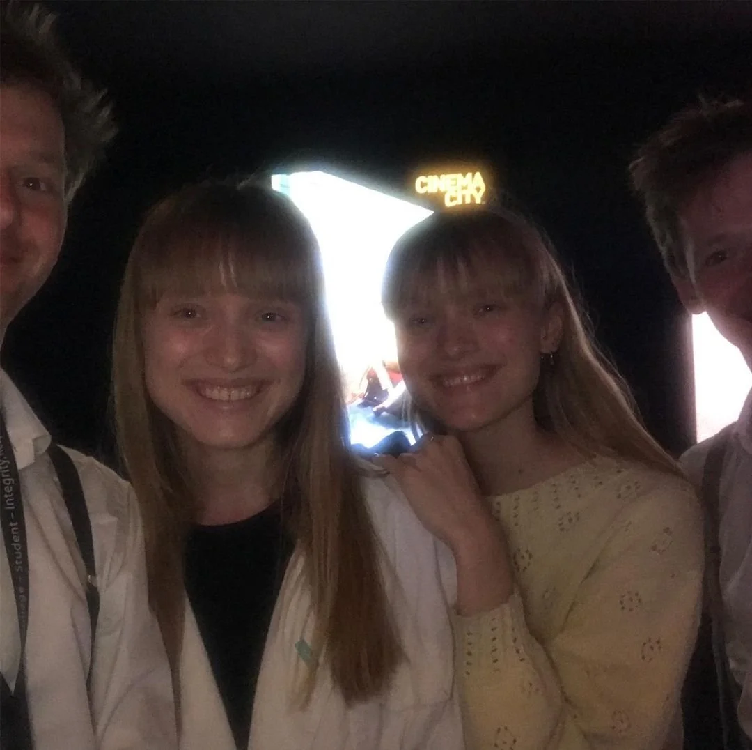 Four young people smiling and posing for a photo in a dark venue with a bright sign that reads 'CINEMAX' in the background.