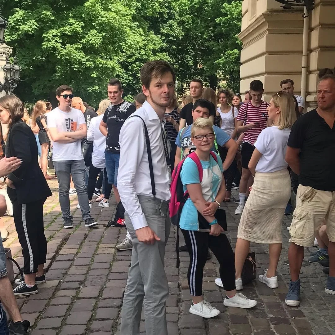 A group of people, including a young girl with glasses and a pink backpack, standing outdoors on a cobblestone pathway with green trees and a building in the background.