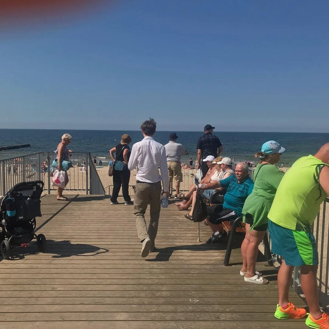 People sitting and standing on a wooden pier overlooking the beach and ocean under a clear blue sky.