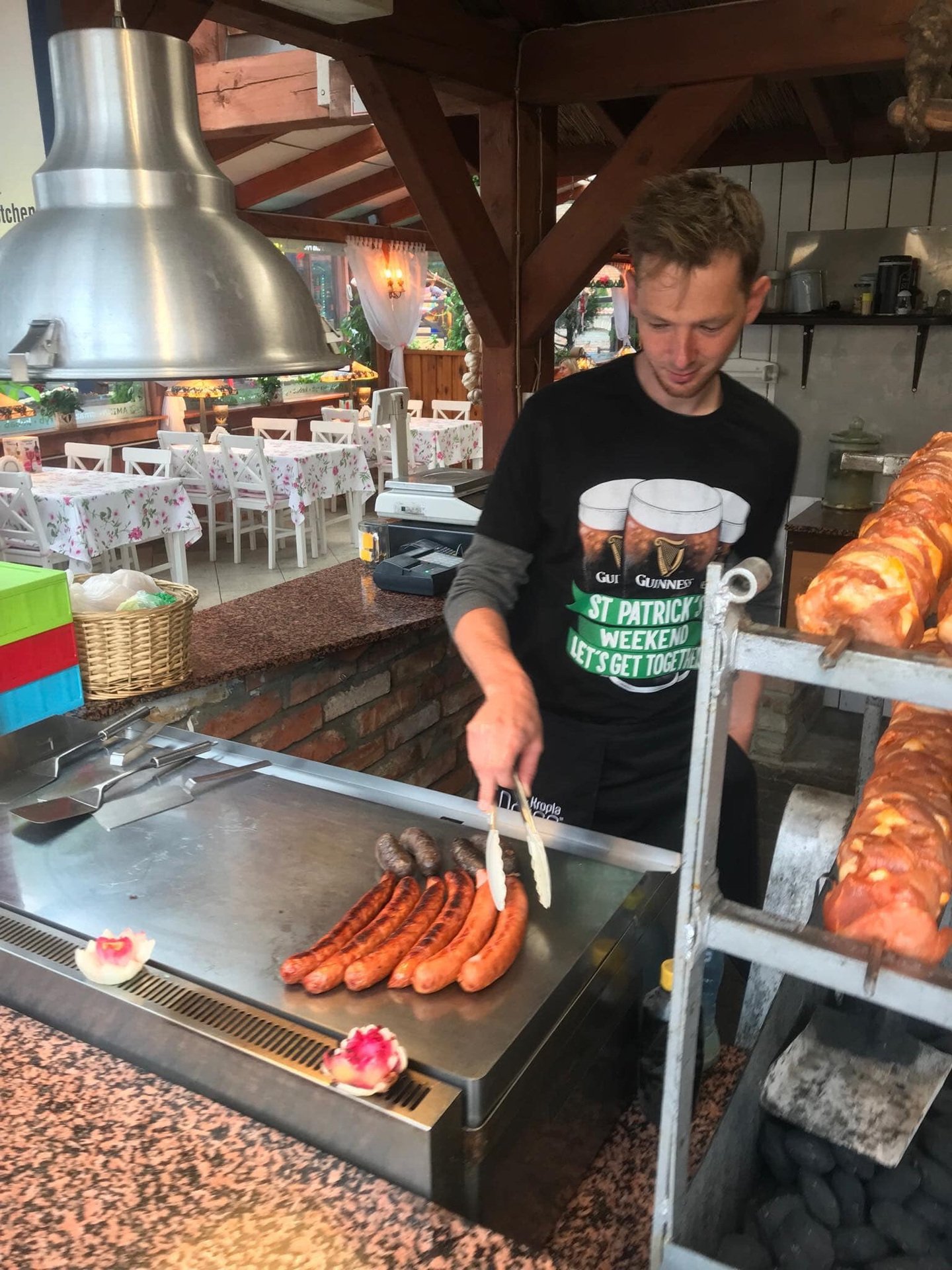 A man cooking sausages and pork skewers at a food stall with an outdoor seating area in the background.
