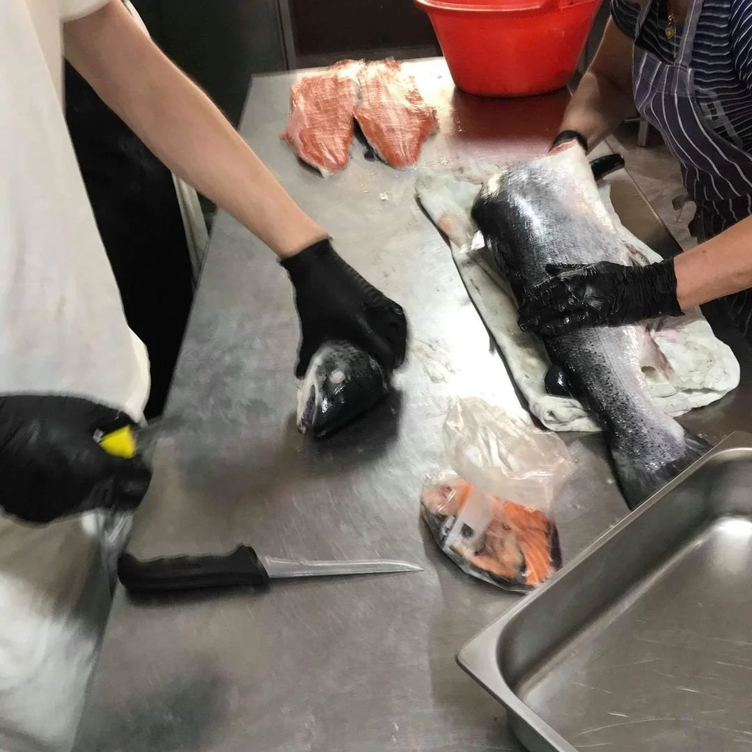 Two people preparing a fish on a metal table, with fish fillet pieces, a whole fish, a knife, and a plastic bag with fish pieces visible.