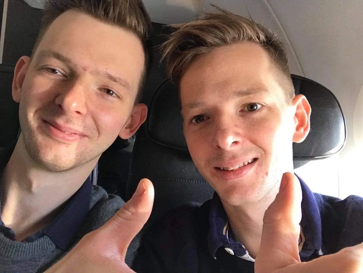 Two young men sitting next to each other on an airplane, smiling with thumbs up, taking a selfie.