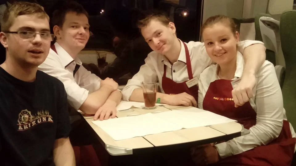 Group of four young people sitting at a restaurant table, three wearing white shirts and red aprons, one in a black shirt, with a window in the background.