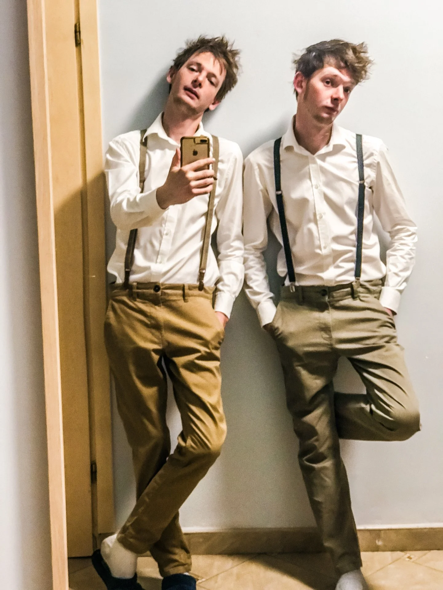 Two young men with messy hair wearing white shirts, khaki pants, and suspenders pose for a mirror selfie against a plain wall. One holds a phone, and both have relaxed expressions.