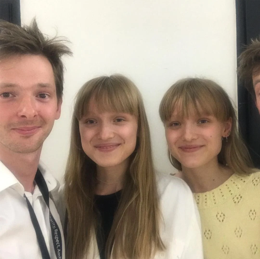 Three people smiling and taking a selfie, two women with blonde hair and one man with brown hair, standing against a plain wall.