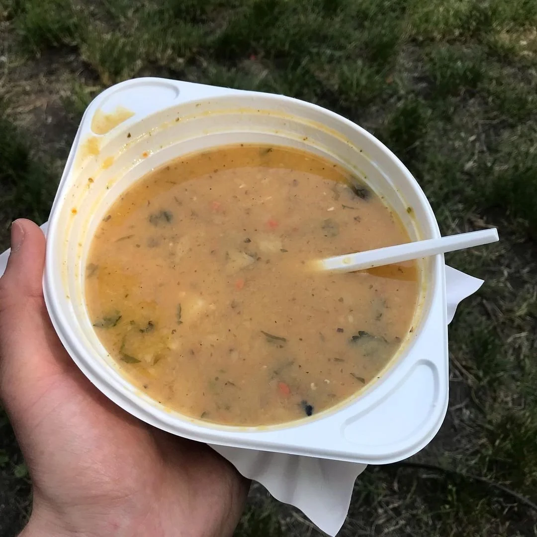 A hand holding a large white bowl of hot, creamy soup with herbs and vegetables, with a white spoon inside, outdoors on a grassy area.
