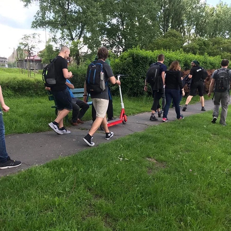 Group of children walking on a park sidewalk with backpacks, one riding a red scooter, surrounded by green grass and trees.
