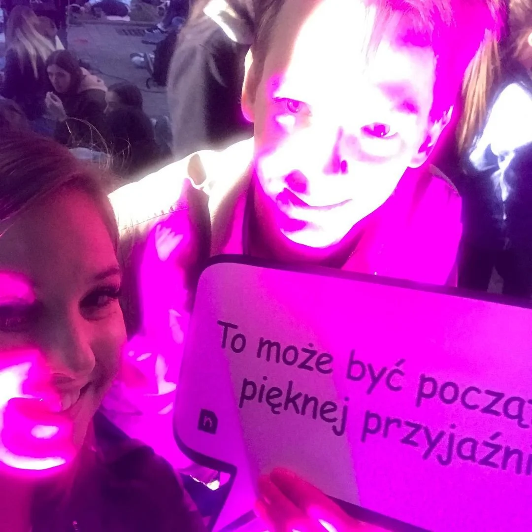Two young people smiling and holding a sign that says 'To może być początek pięknej przyjaźni' with a crowd in the background, illuminated by purple and pink lighting.