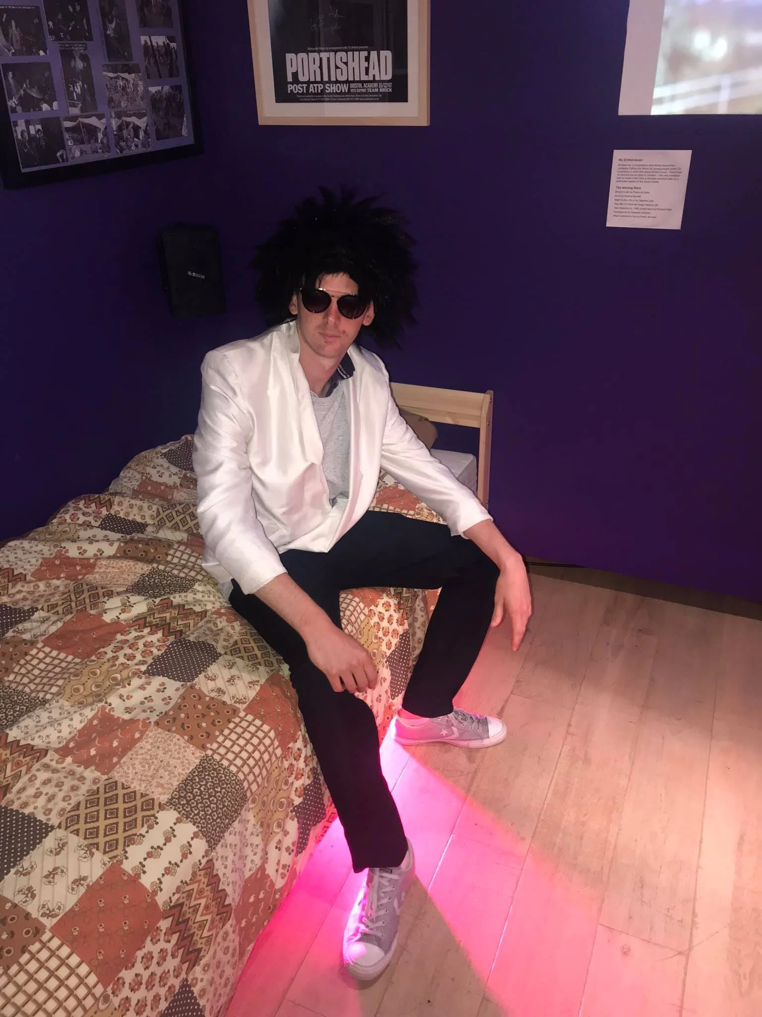 Person dressed as Elvis Presley with black wig, sunglasses, white jacket, sitting on a patterned quilt-covered bed in a room with purple walls, framed posters, and a wooden floor.
