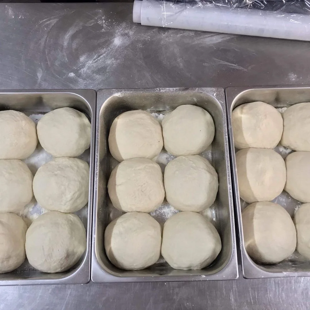 Multiple trays of round dough balls on a stainless steel surface, with a rolling pin at the top of the image.