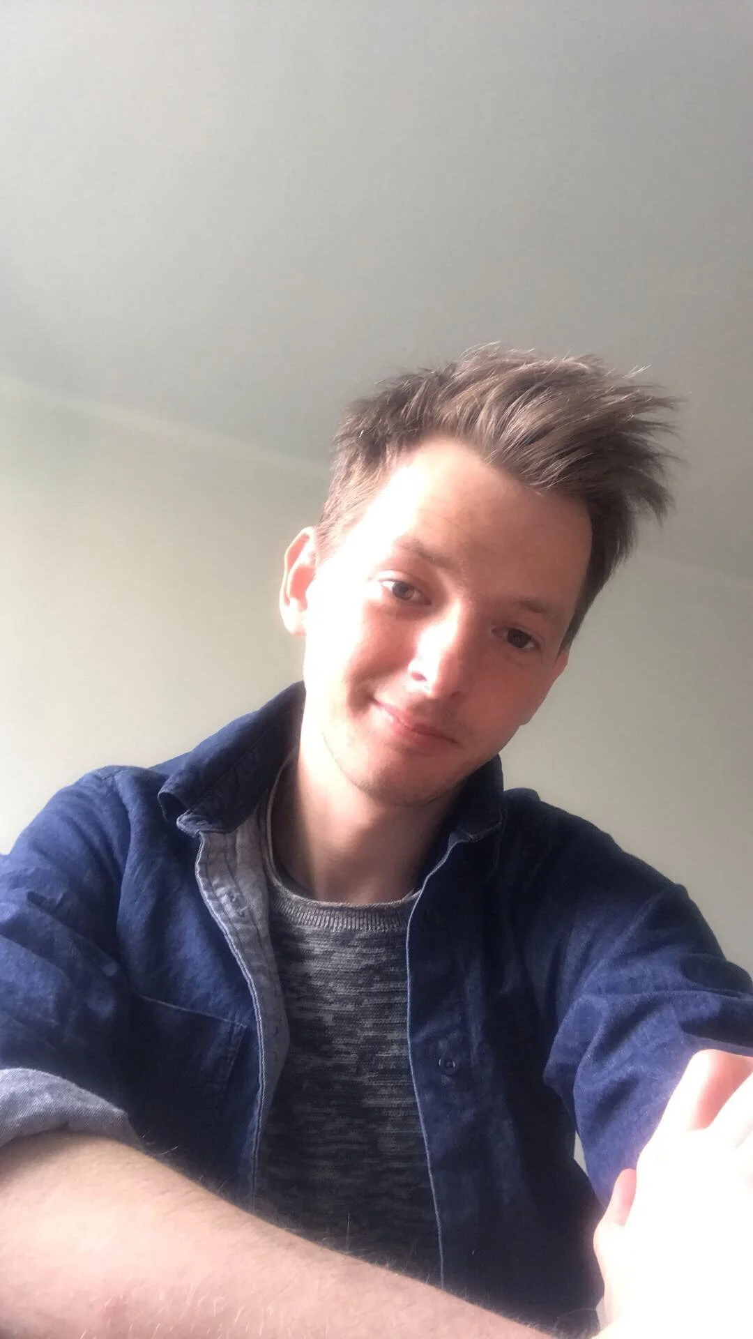 A young man with styled hair, wearing a denim jacket over a striped shirt, taking a selfie with a neutral expression against a plain background.