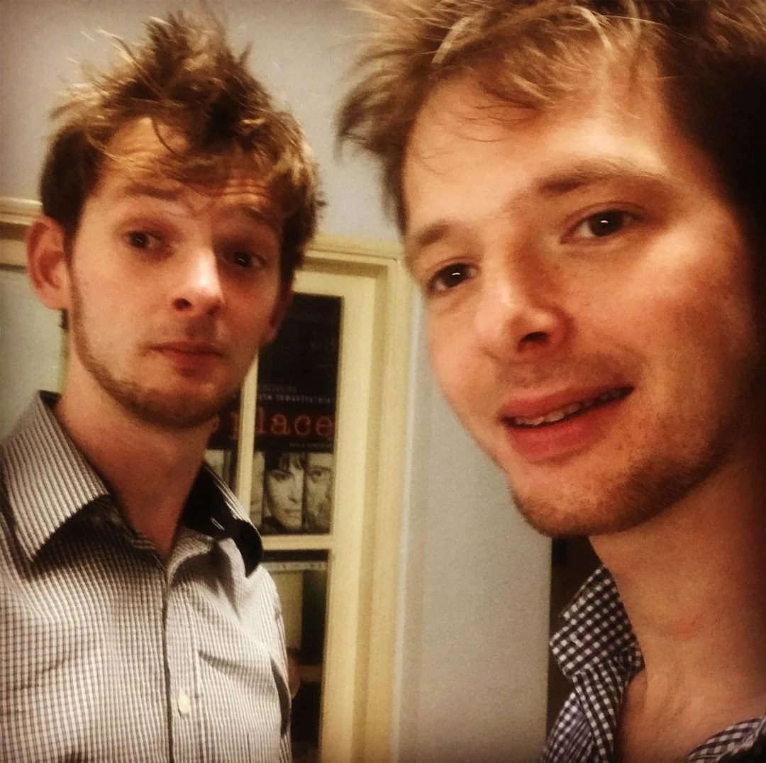 Two young men with messy hair taking a selfie indoors, with a bookshelf or cabinet in the background.