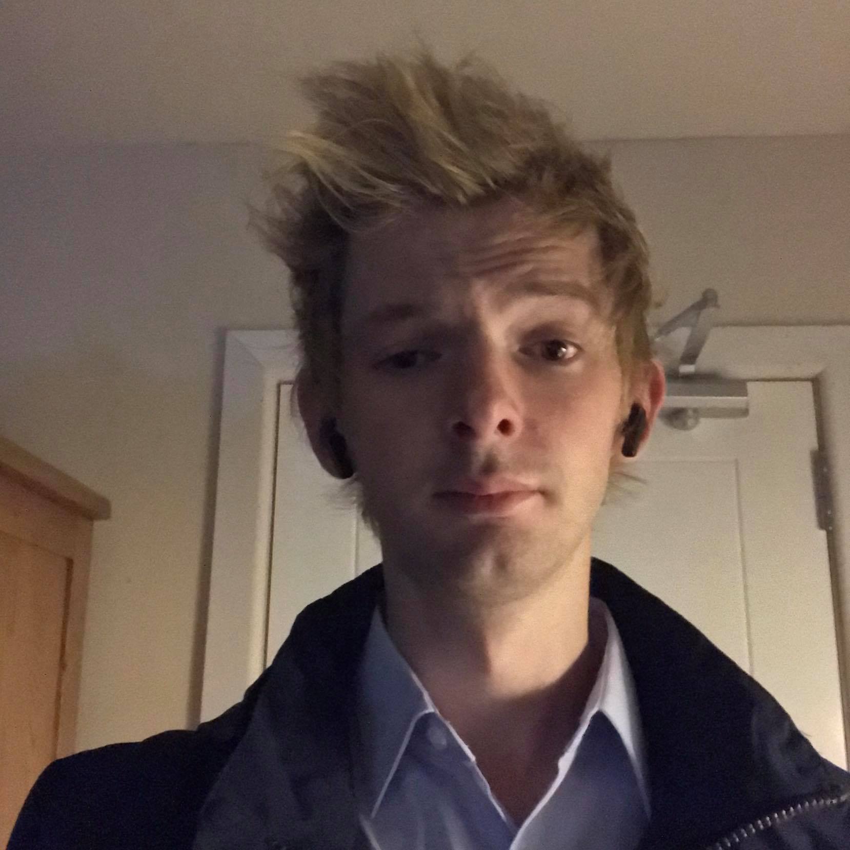 A young man with blonde, messy hair and light skin taking a selfie indoors. He is wearing a collared shirt and a dark jacket, with earbuds in his ears. The background shows a closed door and a wooden shelf.