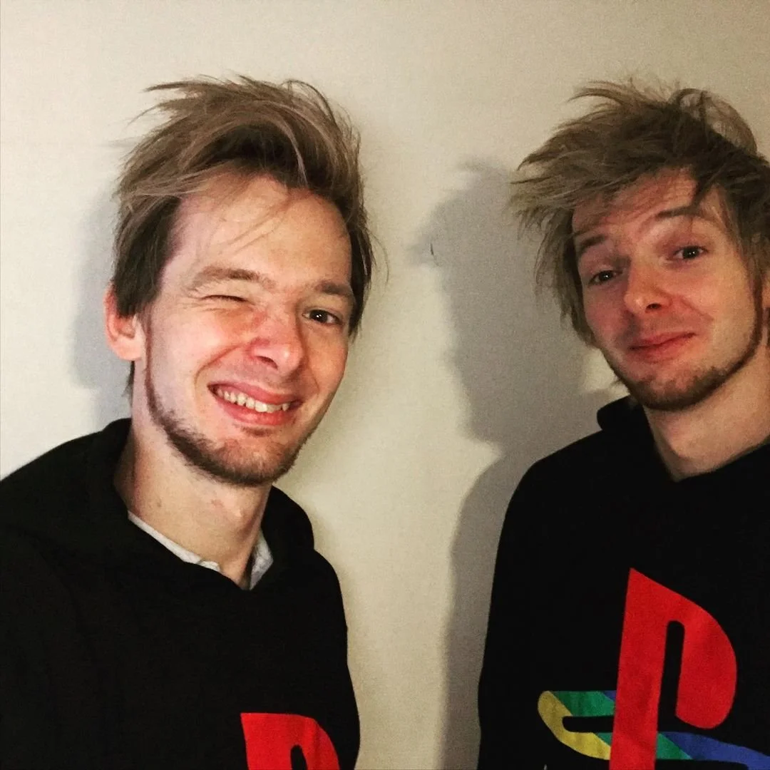 Two young men taking a selfie, both wearing black hoodies with colorful PlayStation logos. The man on the left is winking and smiling, the man on the right has a slight smile. They are standing against a plain white wall.