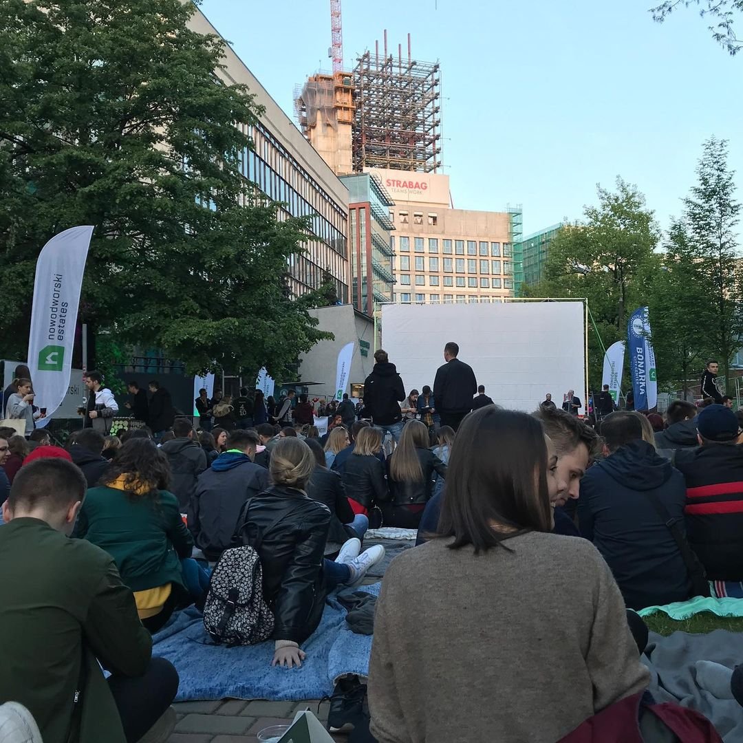 People gathered outdoors on blankets and chairs facing a large blank screen, attending an event in an urban setting with trees, buildings, and a construction site in the background.