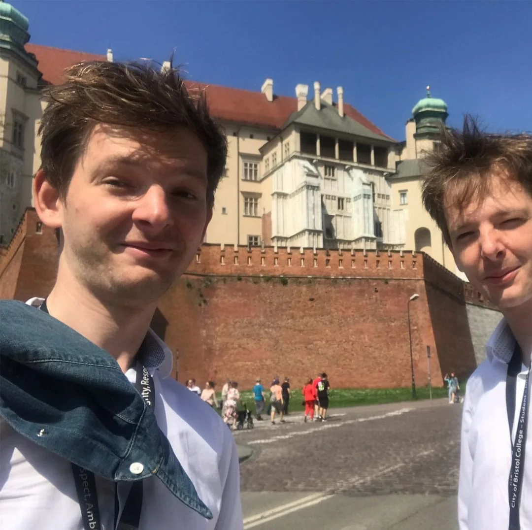 Two young men taking a selfie in front of a historic castle or mansion with a brick wall, under a bright blue sky.
