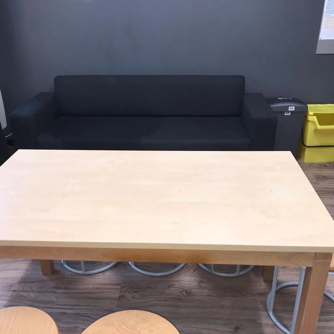 A black couch and a light-colored wooden table in a room with grey wall and wooden floor. There are two yellow containers and a grey electronic device on the right side of the room.