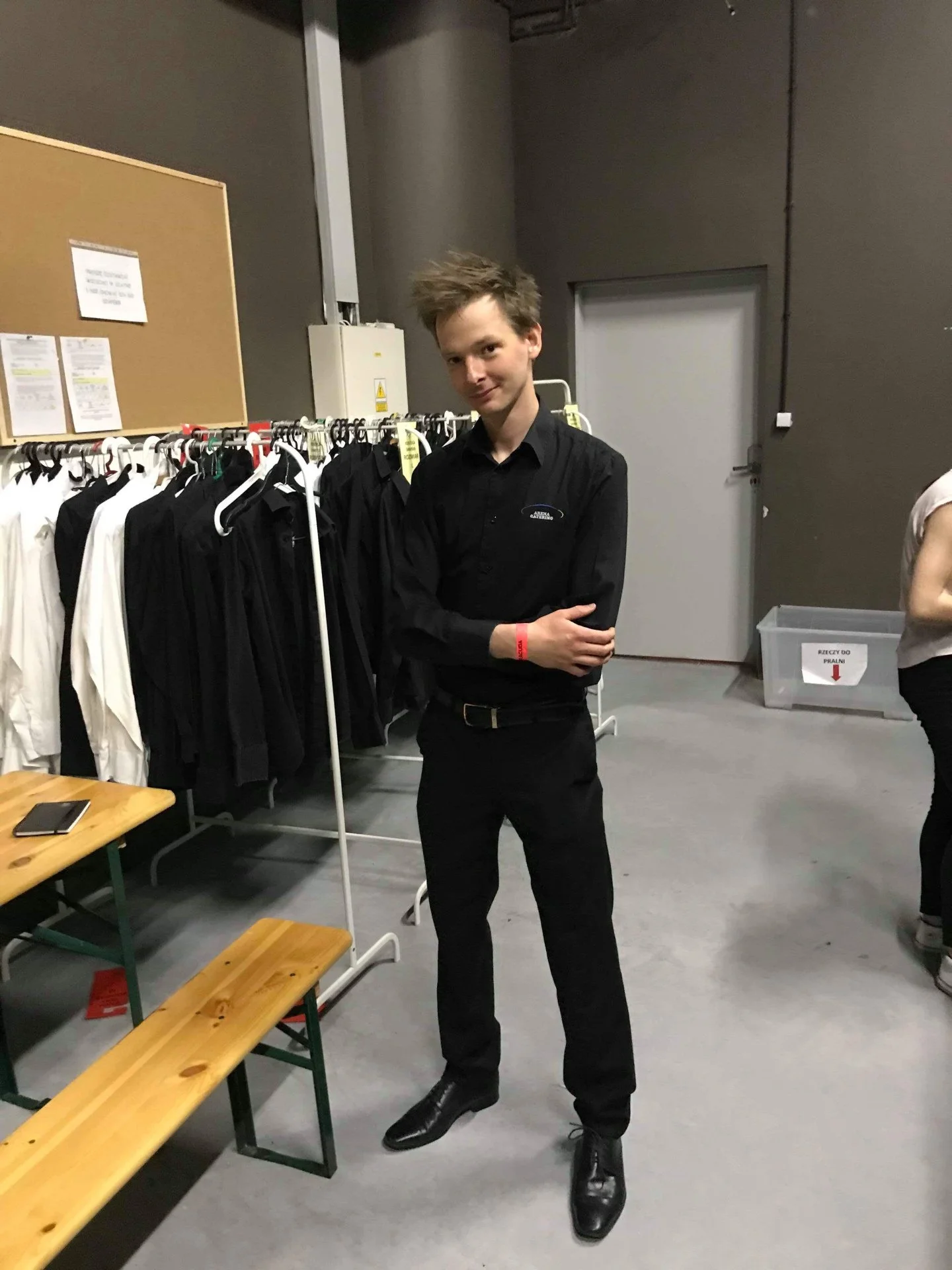 Young man in black shirt and black pants standing in backstage area near clothing rack with black and white shirts, leaning slightly to his left with arms crossed.
